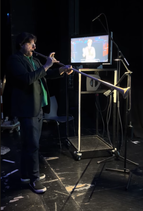 A screenshot of a Youtube video showing a man dressed in a dark jacket and dark pants and a green shirt playing a 5 foot instrument. It is a single long tube with a trumpet-like valve in the middle and ending in a trumpet-like bell, but the entire instrument is made of wood