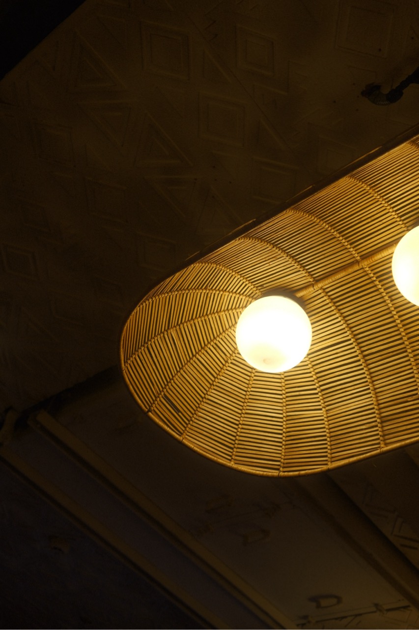A close-up view of a decorative pendant light fixture. The light has a woven, conical shade made of bamboo or rattan, featuring two round, glowing bulbs. The background is dark, highlighting the warm light emitted from the bulbs and the intricate patterns of the shade.