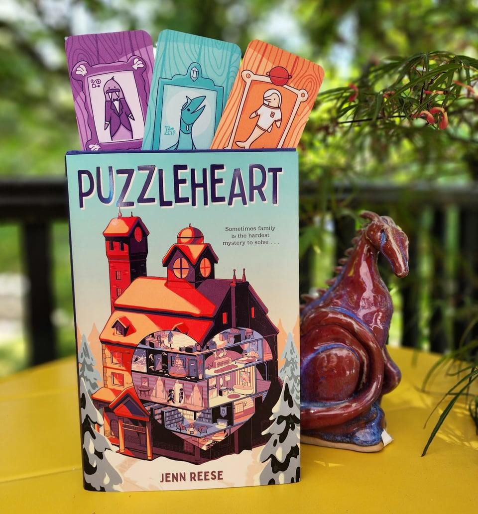 A hardcover copy of my book, Puzzleheart, sits on a bright yellow table under the shade of a tree. A sculpted dragon lurks behind it.