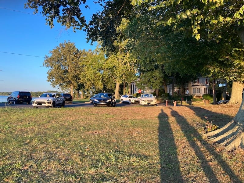 Two people's long afternoon shadows. Cars in background. River to the left. Grass in foreground. 