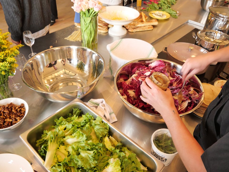 Someone mixes a salad on the stainless steel countertop in the kitchen, surrounded by pots and pans and food in the making.