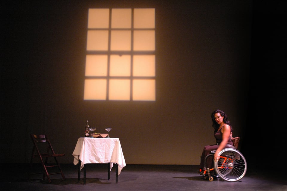 A photo from a Sins Invalid performance. On a softly lit stage, a person with medium brown skin, long wavy hair, and wearing a dark brown sleeveless dress rolls her wheelchair up to a table. The table is simply but elegantly set for two, and an empty chair waits for a companion to join the woman.