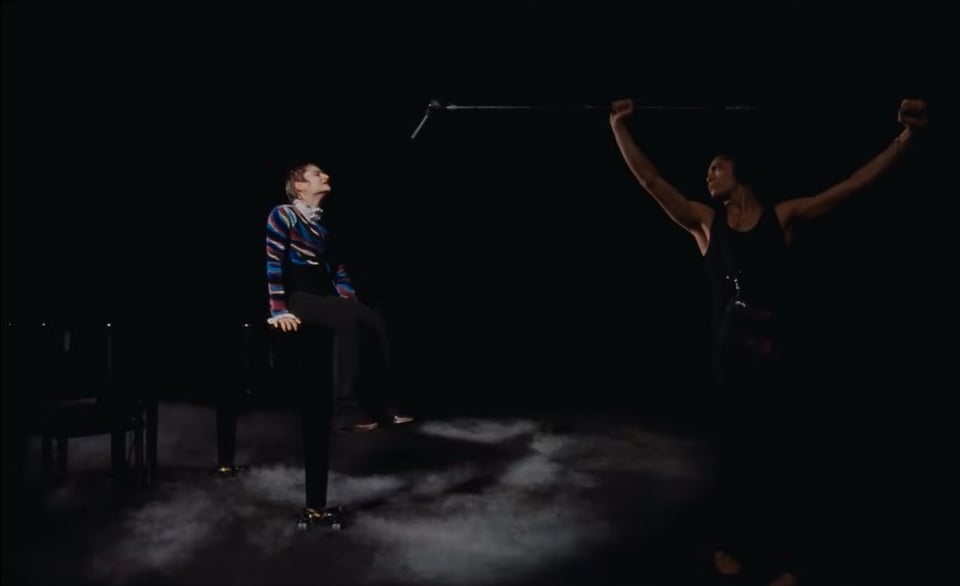 A still from the video for Christine And The Queens' 'Mountains' featuring Christine sitting on a piano in front of a dancer holding a microphone on a boom pole.