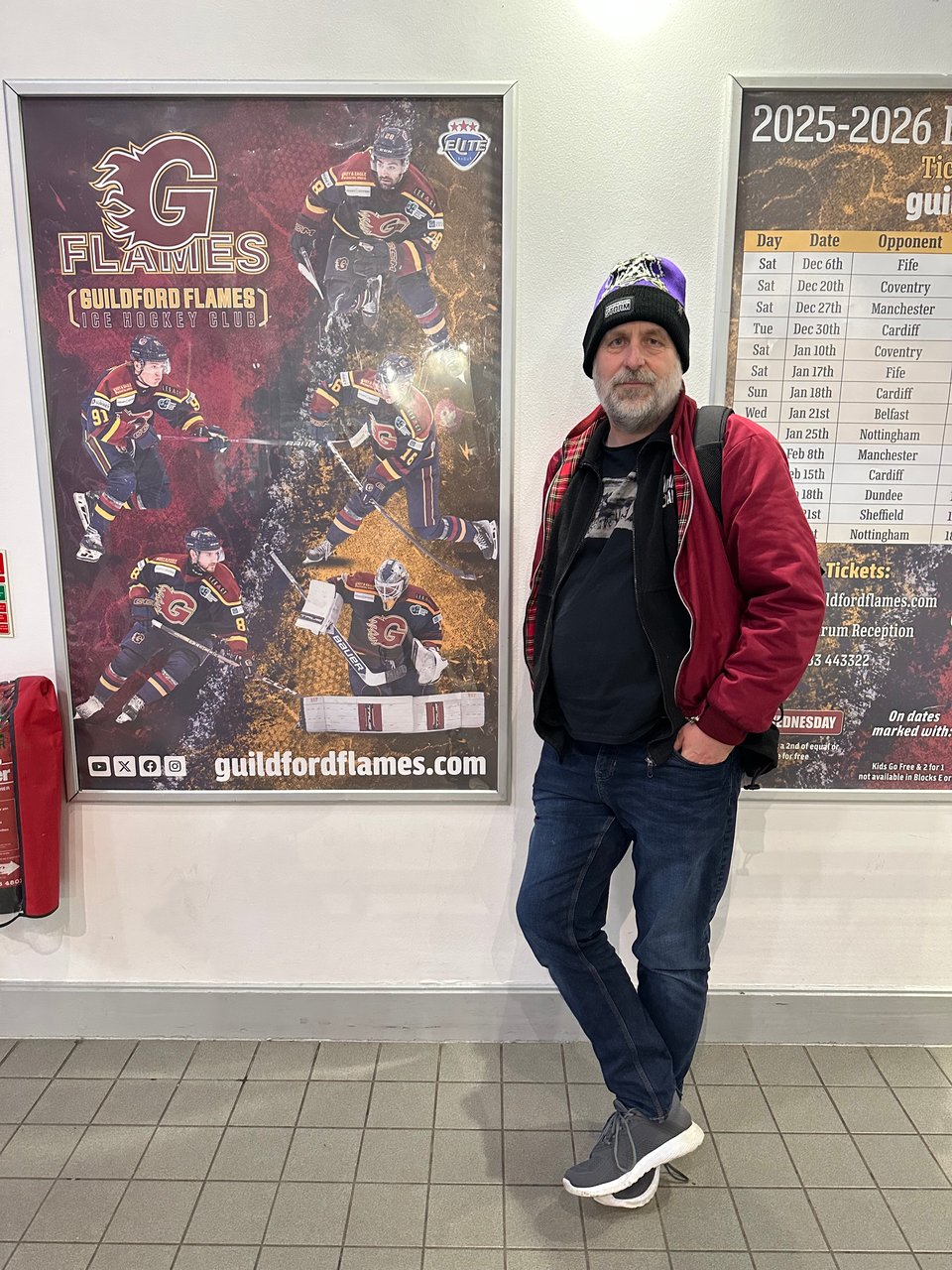A man standing on a neat tiled floor before a beige wall, upon which are two posters advertising Guildford Flames.