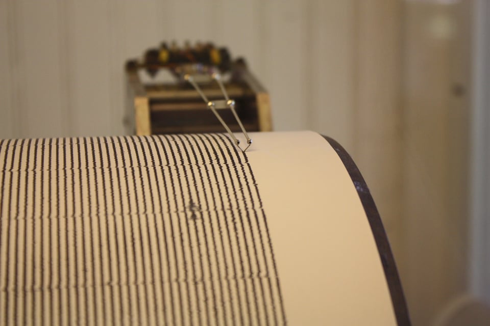 A close-up view of a seismograph machine shows a needle drawing fine, wavy black lines across a rotating drum of paper. The steady, parallel traces indicate low seismic activity, with the instrument poised to record any vibrations or tremors. The background is softly blurred, focusing attention on the precision of the recording mechanism.