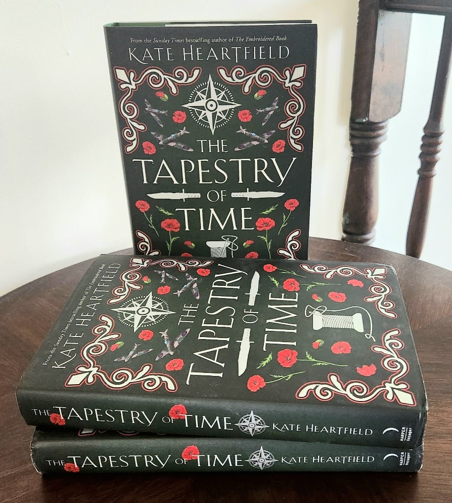 Two hardcovers lie horizontal and one stands upright behind it on a table. The cover is very dark green with a compass rose, a spool and poppies surrounding the title, The Tapestry of Time.