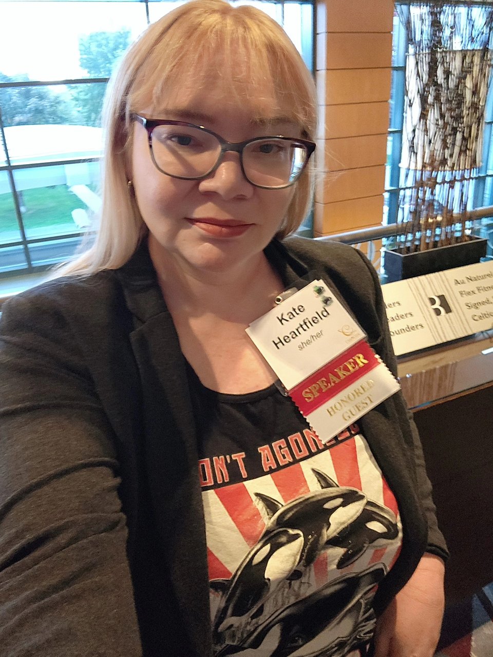 A selfie of Kate, a middle aged white woman with long blond hair, wearing a grey suit jacket over a tank top that says "Don't Agonize, Orcanize" and a convention badge that says "speaker" and "honoured guest."