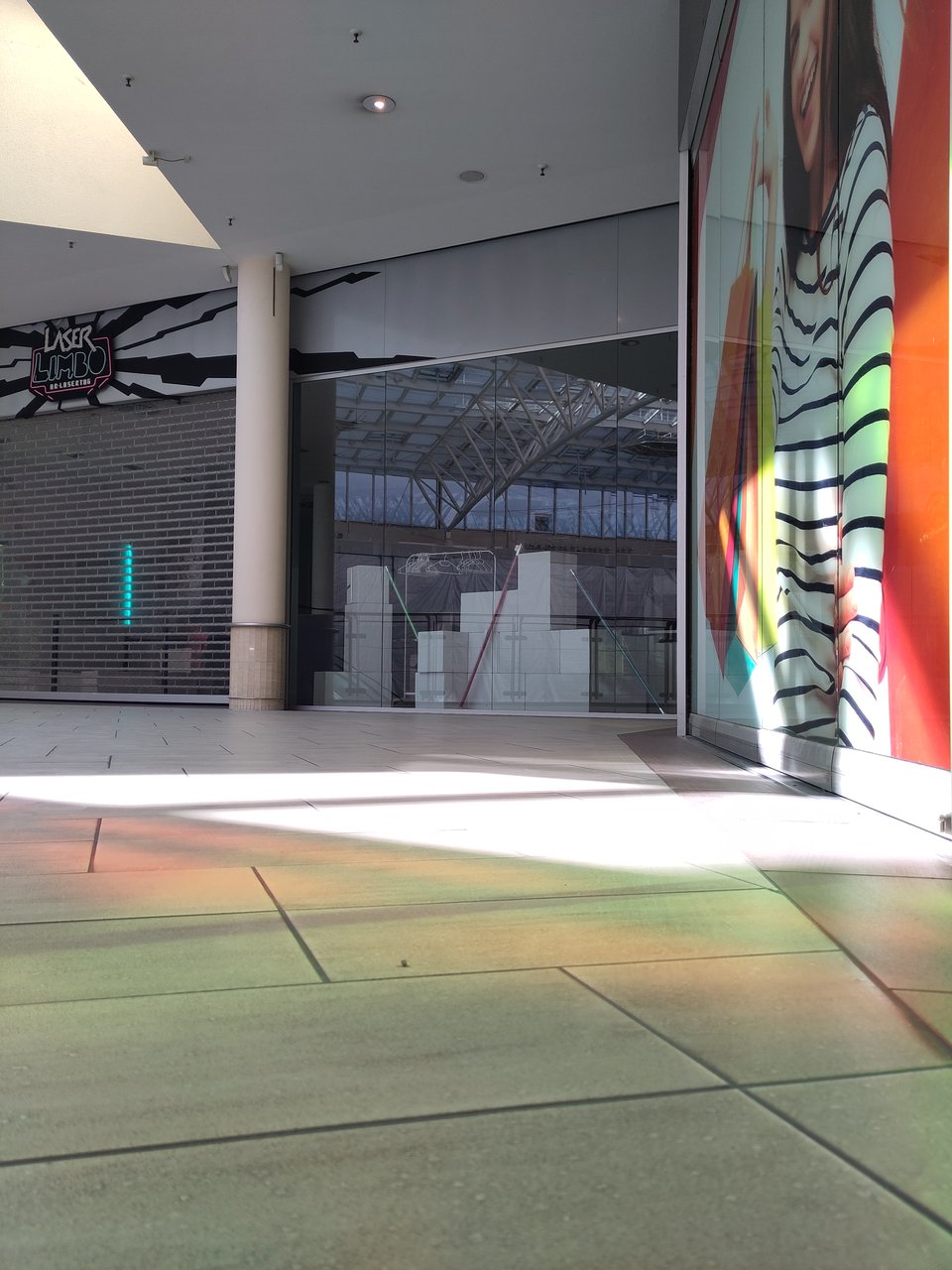 Ausschnitt einer dem Anschein nach leeren Ladenpassage. Auf dem Boden großflächige Fließen, zwei Ladenlokale geschlossen, eines im Schaufenster mit unbestimmbarer unmotiviert wirkender Deko.