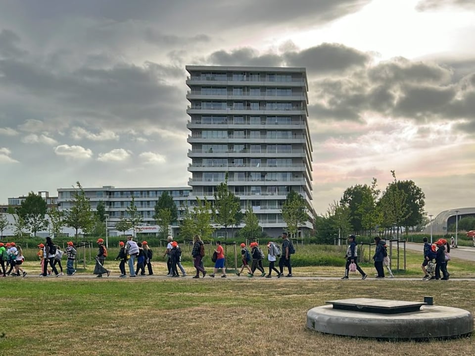 Children walking in a line through a park with a high rise apartment building in the background.
