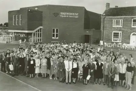 A large group of people standing in front of a newly opened theatre. The building in the background is brick, with a faintly hexagonal form. On the front wall a large block overhangs the entrance steps. I feel like I'm getting a static charge from all the nylon.