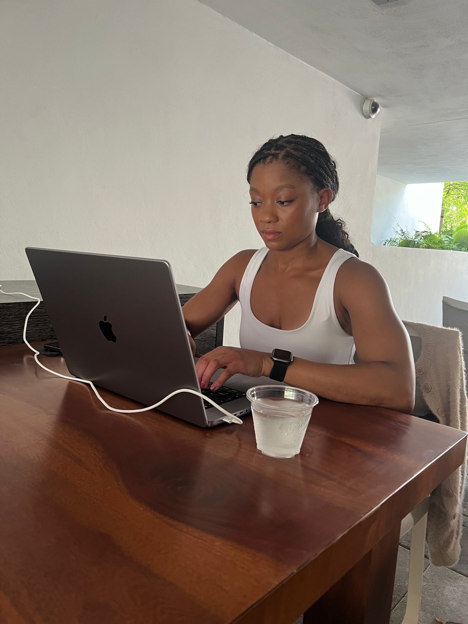 Image of a me working on a laptop at an outdoor setting. I'm waring a white tank top, working on my MacBook at a wooden table. I'm wearing a watch and have braided hair, with a glass of water beside me.