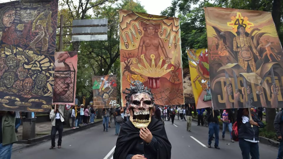 A street poster features elaborate banners with indigenous imagery and a man wearing a skeletal mask