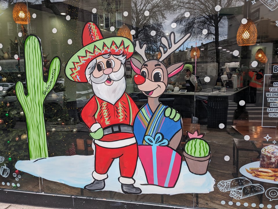 A hand-painted decal on a window featuring (left to right) a cactus, Santa Claus wearing a red suit and a red and green sombrero, a reindeer (atop a boxed present with bow) wearing a blue serape, and a smaller plant.