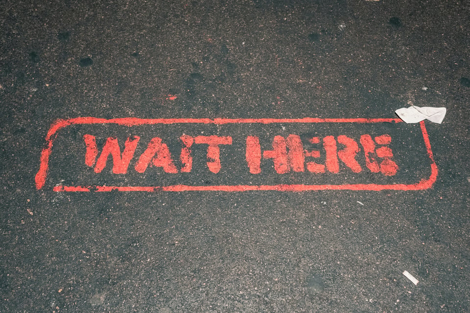 Photo of a section of black pavement or asphalt with the words "Wait Here" in red paint, surrounded by a red border stamped on it.