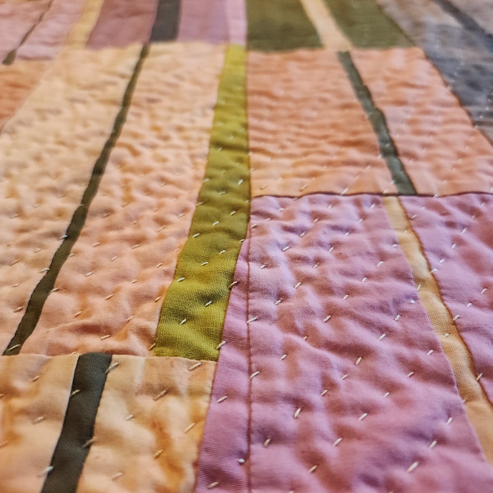 Close up photo of Arabesque, a patchwork quilt by Sarah Atlee, with fabrics in peach, pink, purple, and olive green. Sarah’s hand stitches are visible.