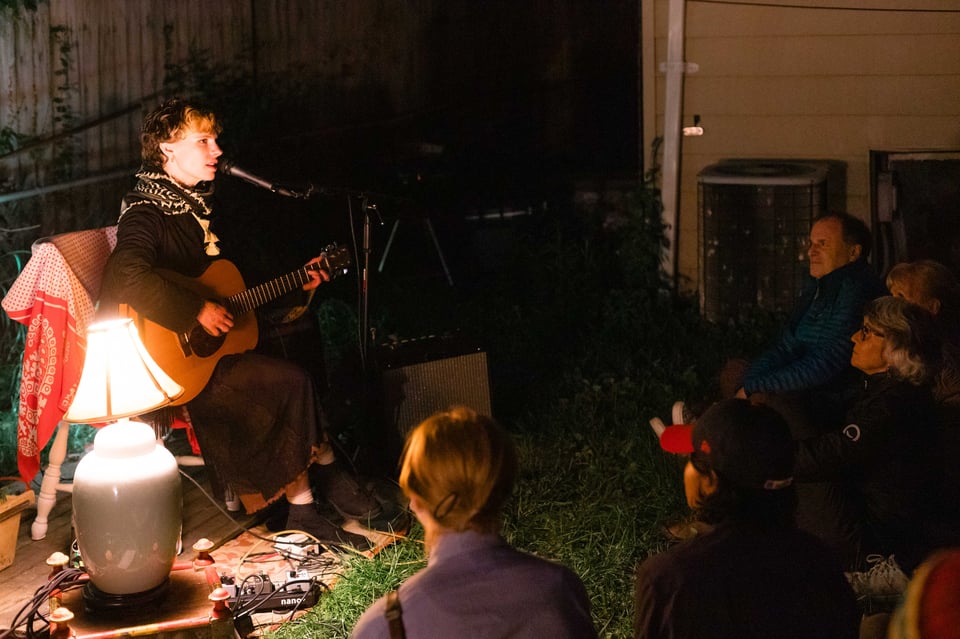 Tallen Gabriel is on the left of the image, lit by a lamp, wearing a keffiyeh, playing guitar to a crowd sitting on the grass.