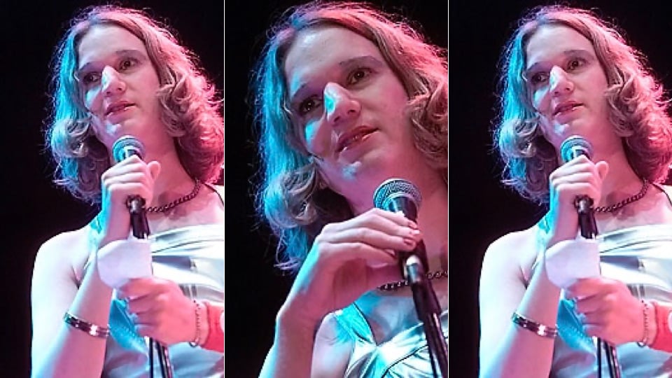 A trans woman with long blond hair wearing a chain necklace and bronze bracelet, plus a silver lame dress, reading off a scrap of paper into a microphone.