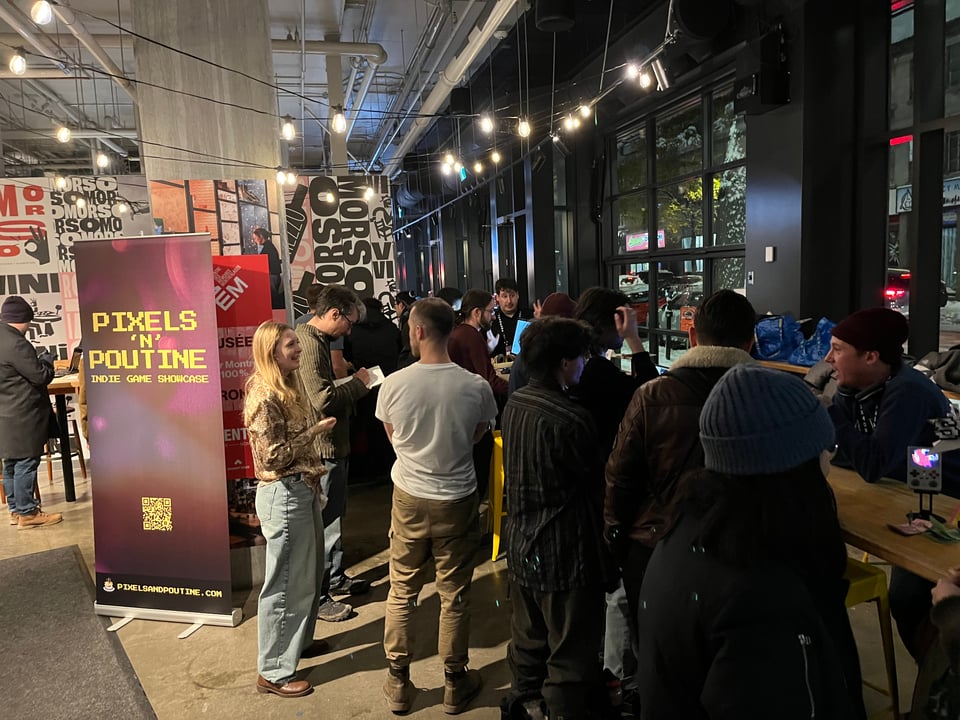 A group of people stand around a table and chat. One person is playing a game on a computer. There's a poster that says "Pixels 'n' Poutine Indie Game Showcase."