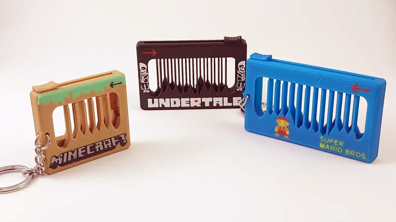 A photo of three of the musical fidget toys. They are small and rectangular, and each one has thick plastic around the edges, and an empty center bisected by perhaps a dozen thin plastic strips. The one on the far left has "Minecraft" written on it in the Minecraft font; the one in the middle has "undertail" written on it in that font; and the one on the right has a small icon of Mario on it and the text "super Mario Brothers"