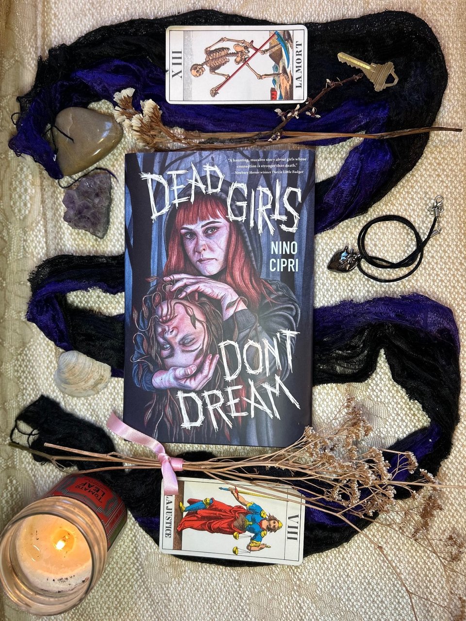 The book Dead Girls Don't Dream in a flat overlay. It's surrounded by witchy accessories: dried flowers, rocks, shells, a lit candle, a key, and a charm shaped like an anatomical heart. At the top of the book is the tarot card of Death. At the bottom is the tarot card of Justice.