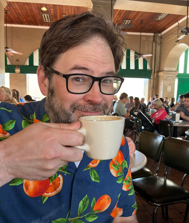 This is a photograph of me sitting in the Cafe Du Monde in New Orleans. I am wearing a blue shirt with bright oranges on it. I look like a dad on vacation. I am holding up a cup of coffee as I look at the photographer. I smile in a way that I can only smile when I am on vacation, away from the stresses and worries of everyday life.