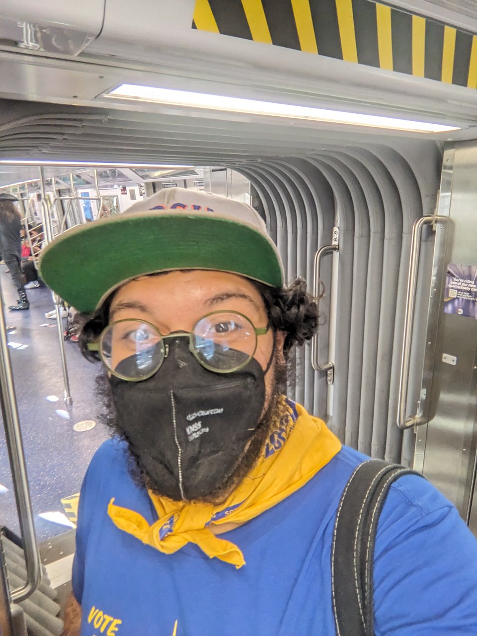 the author wearing a blue shirt, yellow bandana, black covid mask, glasses with dark rims, and a gray hat with a green bill, standing in the open gangway of a G train.