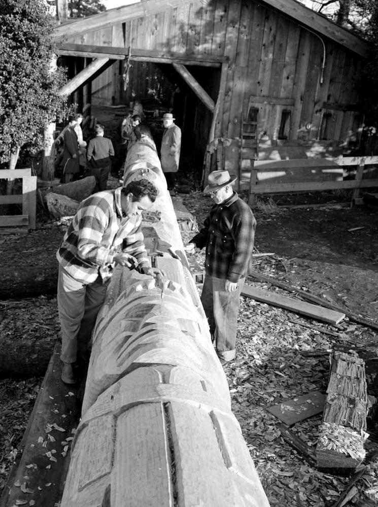 Black and white photo of two men carving a totem