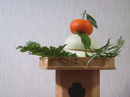 Kagami mochi with an orange tangerine sitting atop two pieces of white mochi perched with some greenery on a wooden stand.