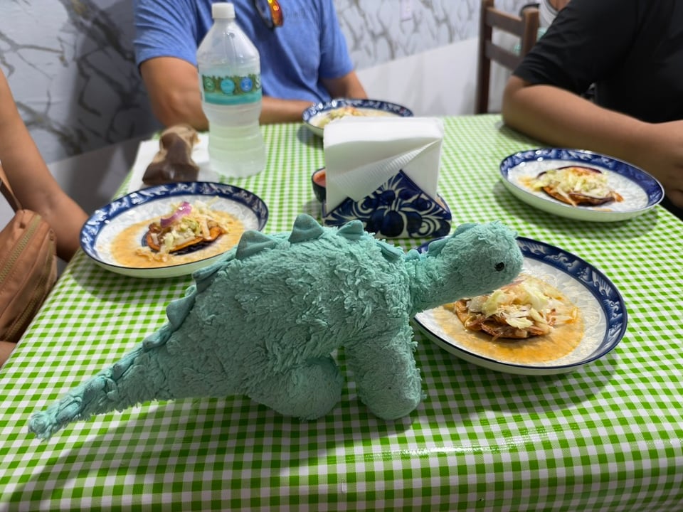 Chomp, a stuffed blue-green dinosaur, eating a Mexican sopito on a vinyl-covered restaurant table. His beady eyes hide a dark purpose.