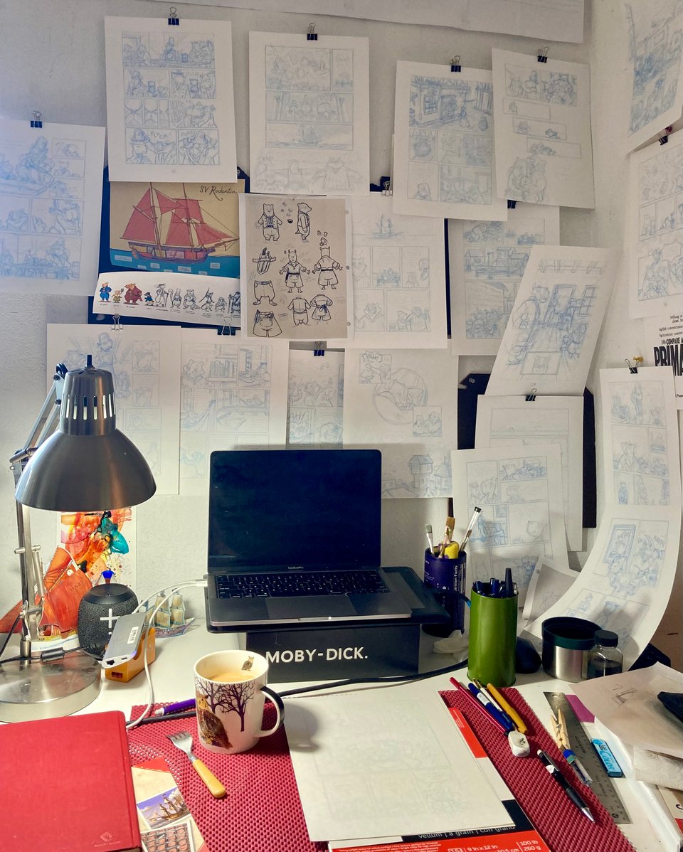 Lucy's cluttered desk with loads of penciled comics pages hanging from the walls around it. Her laptop sits on a copy of Moby-Dick.