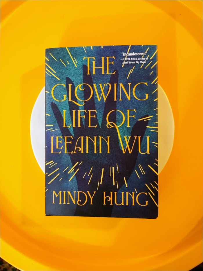 A flatlay of Mindy Hung's THE GLOWING LIFE OF LEEANN WU on a white circle (it's a trivet) and in a yellow circle (my coffee table)