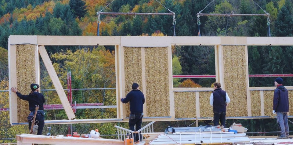 Großes Holz-Stroh-Lehmelement schwebt an Trägern und wird von vier Personen montiert.