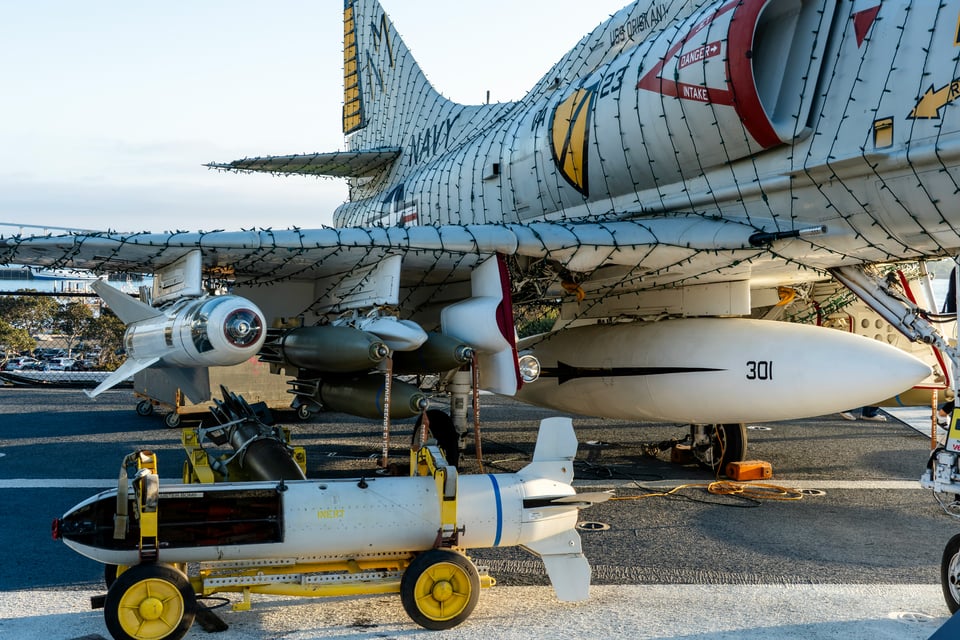 An A4 Skyhawk sits wrapped in Christmas lights at the USS Midway Museum, in San Diego, CA. You can see the tail and wing of the jet, with weapons mounted under the wing and an example bomb sitting in the foreground, with the cast cut open so you can partially see inside of it.