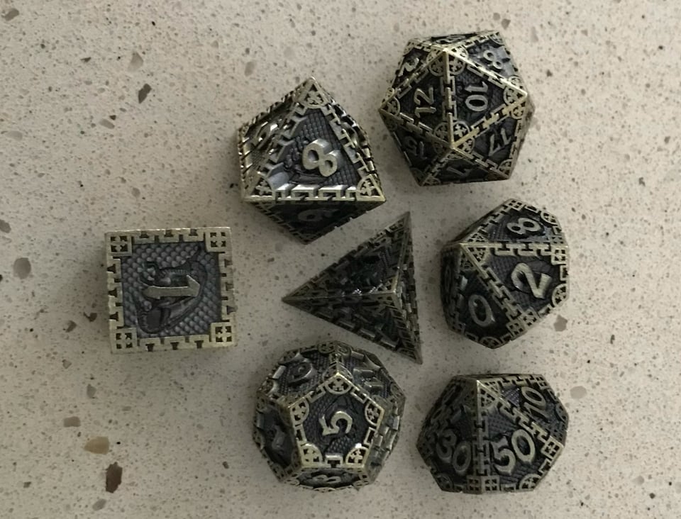 Seven polyhedral dice. They are made of a grey material that looks metallic, with gold numbers and edge trim.