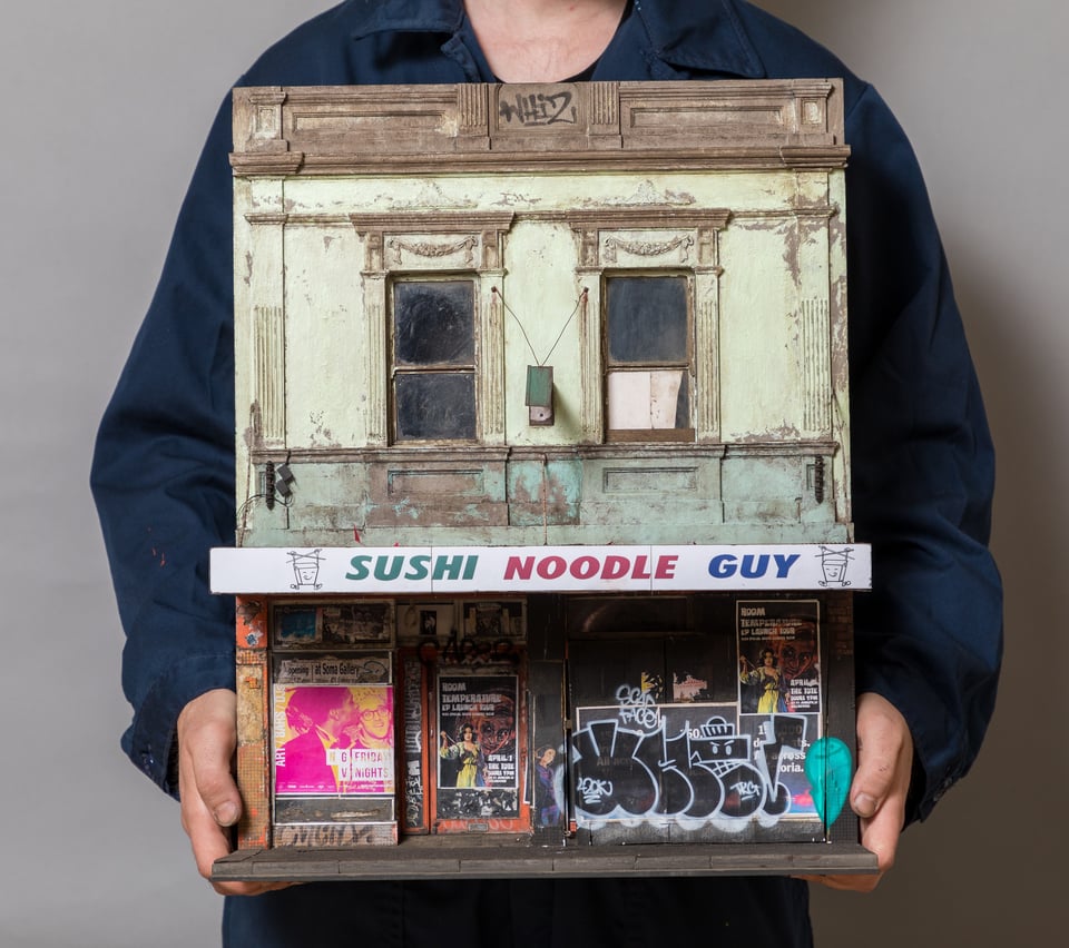 A photo of a man in a blue shirt holding the scale model of a two story building. The building is of a slightly decrepit business that has been shut down and covered with graffiti. That business is called "sushi noodle guy". The graffiti includes a black and white set of icons that look somewhat like numbers, and several posters. The second floor of the building looks like a facade from the 1920s, covered in stained green paint. The photo is cropped such that you cannot see the head of the man.
