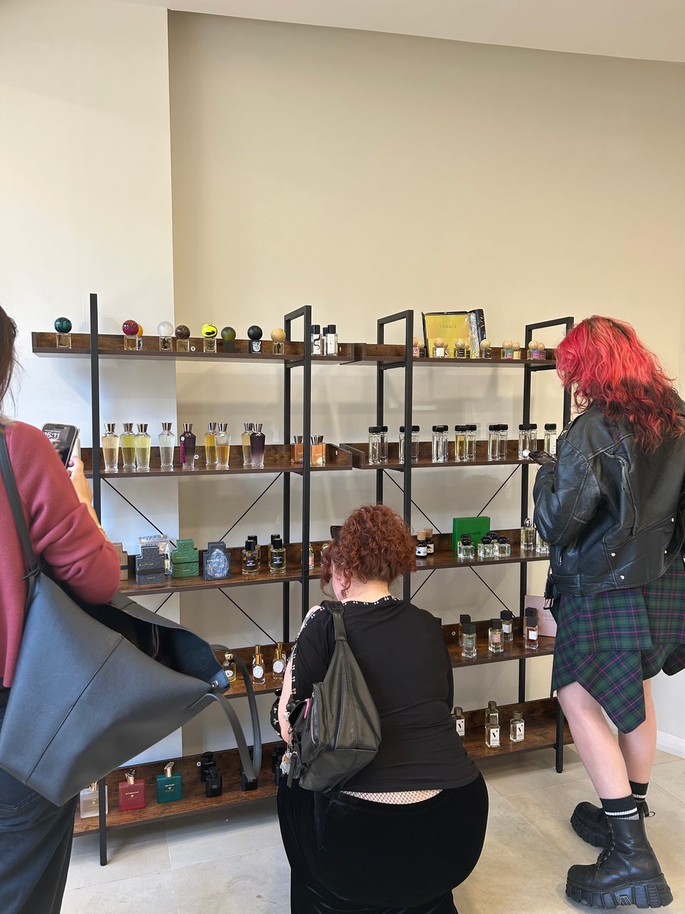 Three people with their backs to the camera are looking at bottles of perfumes from independent brands, displayed on wooden shelving units in a brightly lit space. Image by Rowan Ambrose.
