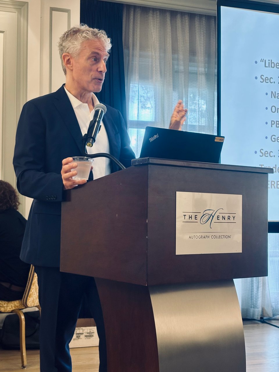 photo of middle-aged white man in navy suit and white polo shirt standing at lectern, holding a plastic cup of water in his right hand and gesturing with his left; there's a mic attached to the lectern and a laptop in front of him.