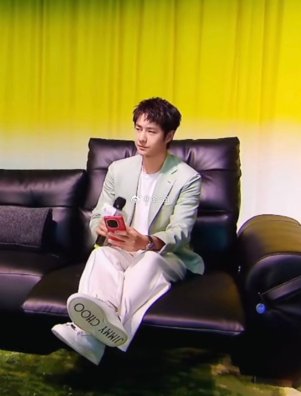Wang Yibo sitting on a brownish-purple sectional with his feet up on a reclining section, wearing a green blazer over a white top and white pants. The soles of his white shoes read, loudly, JIMMY CHOO. He's holding a red phone in one hand and a microphone in the other. His hair is tousled and perfect.