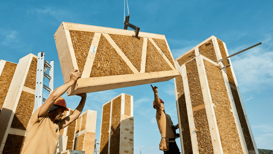 A photo of two workmen reaching up to grab a large wooden structure as it is lowered on a crane towards them. The wooden structure is a rectangular wood frame, the inside of which is stuffed tightly with brown straw. All around them are several similar tall wooden rectangles, standing upright on the ground, also packed full of straw. The sky above them is blue.