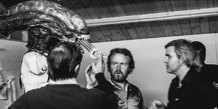 Someone offering a cigarette to a xenomorph’s small inner mouth on set of one Alien. Ridley Scott and H.R. Giger both look on in consternation.