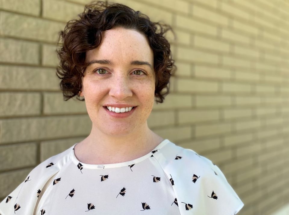 A picture of a white woman with wavy black hair, in a white blouse standing in a hallway