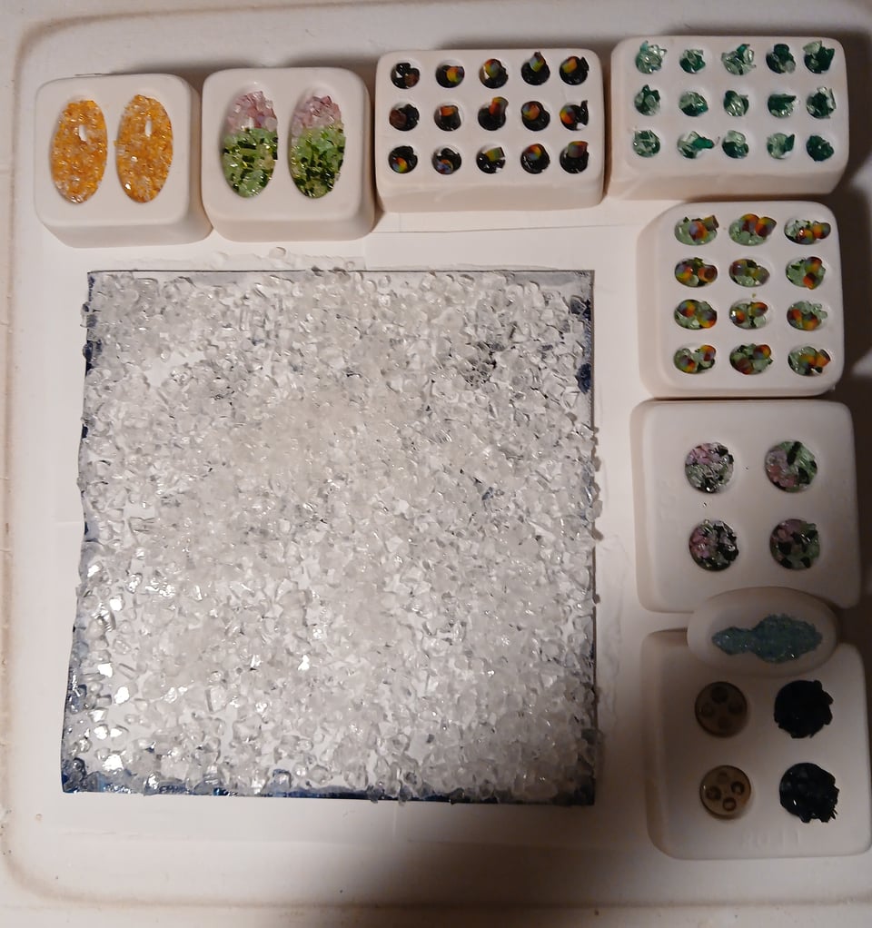 kiln shelf with a big blue sheet covered in white and clear frit and assorted molds full of frit. the most interesting color scheme is a pink and green one. one of the molds is in the shape of a weirdly realistic mushroom