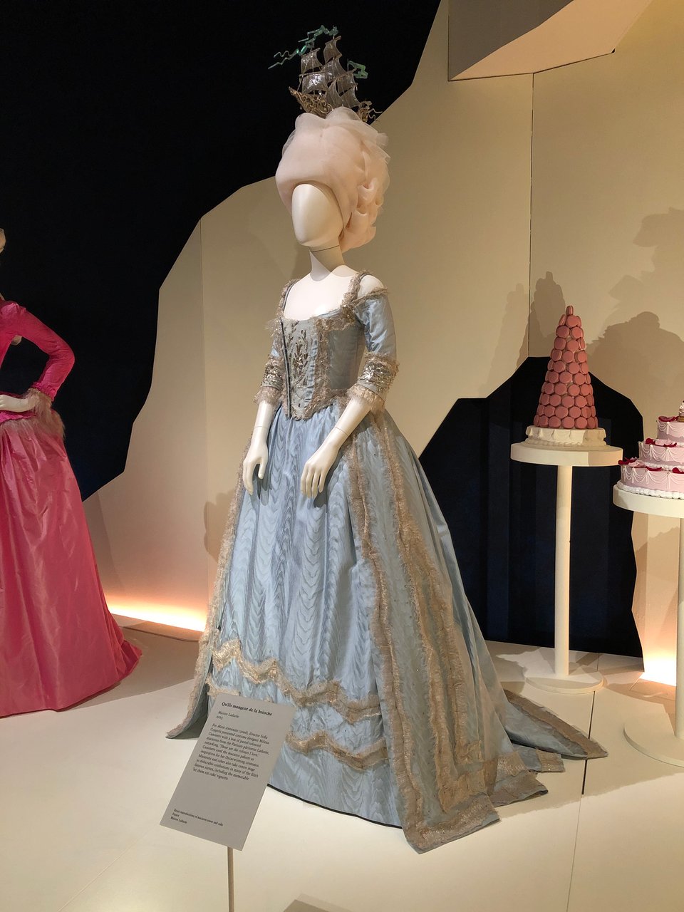 A museum display of a long-skirted, pale blue dress with soft gold details, on a cream-coloured tailor’s dummy. On the dummy’s head is a creamy wig topped with a ridiculous ornament shaped like a sailing-ship. In the background are towers of pink cakes and macarons, and part of a full-length pink dress.