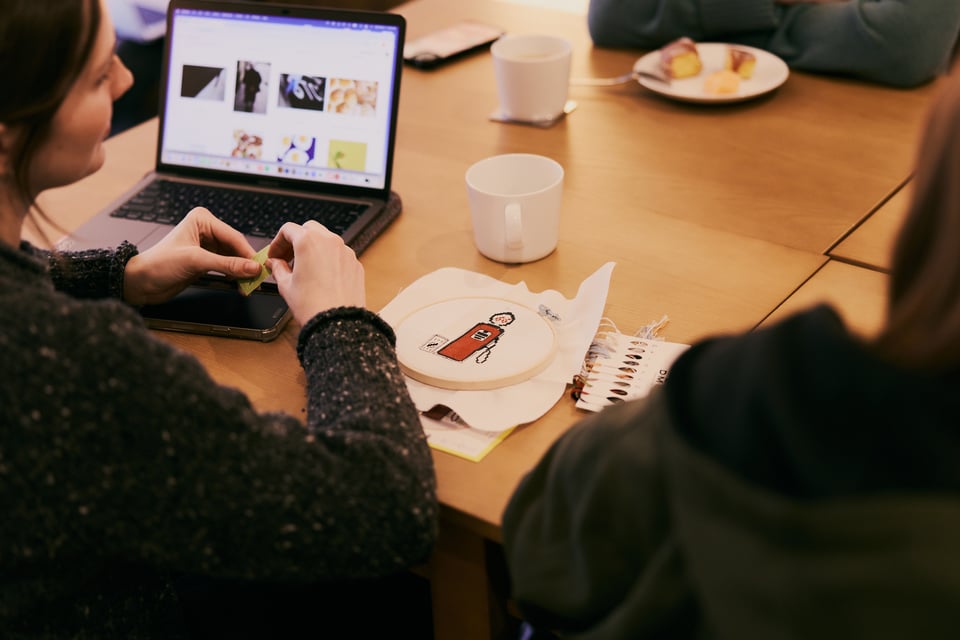 People gathered around a table with pastries, needlepoint, and a laptop.