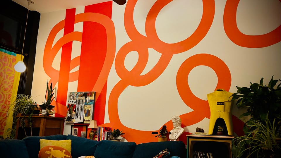 a blue couch with yellow pillows, an old console tv converted to hold records, various artworks on the counter, plants, and a white wall painted with two orange pillars and large lighter orange swirls that match the fabric of a yellow & orange curtain