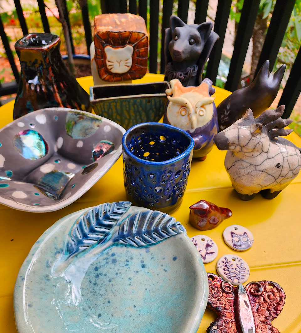 A smattering of ceramics arrayed on a bright yellow table. There are bowls with feathers, some with dots. There is a crackled chonkly white horse, a lion, some weird sort of fox thing. A butterfly and a small bird sit up front in glittering red.