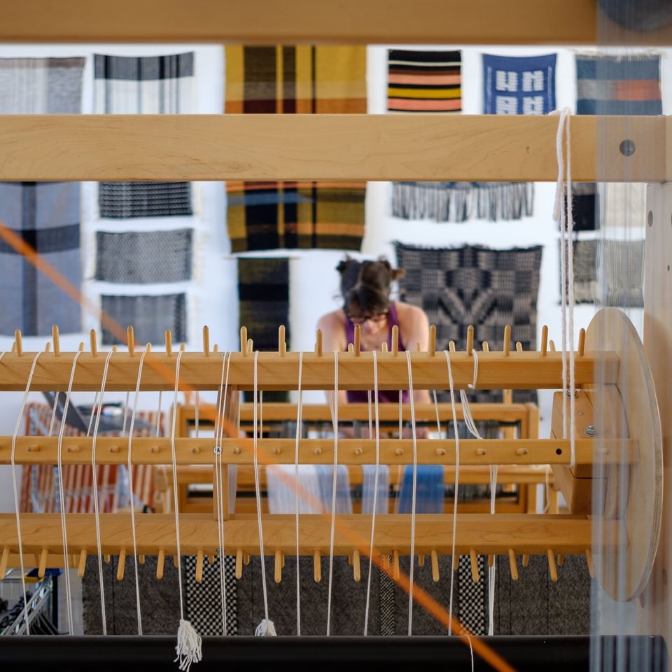 An artist is seen working on a loom. The loom is in focus in the foreground with the artist out of focus in the background. A wall of textiles hang on a wall behind her.