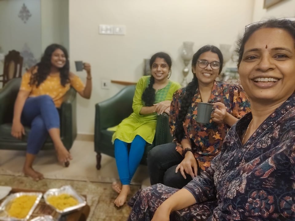 Snacks are placed on a table as four people smile and laugh toward the camera; the person in front is taking a selfie.