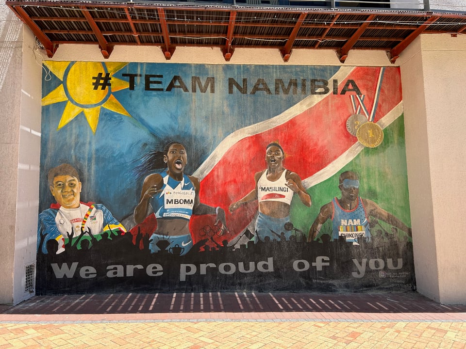 A mural showing four people. Far left is a white man with his arms crossed, smiling. To the right of him is a black woman running with her mouth open and the name MBOMA on her shirt. To her right is another black woman running, with the name MASILINGI on her shirt. And to the far right is a black man wearing an eye covering. At the top it says #TEAM NAMIBIA