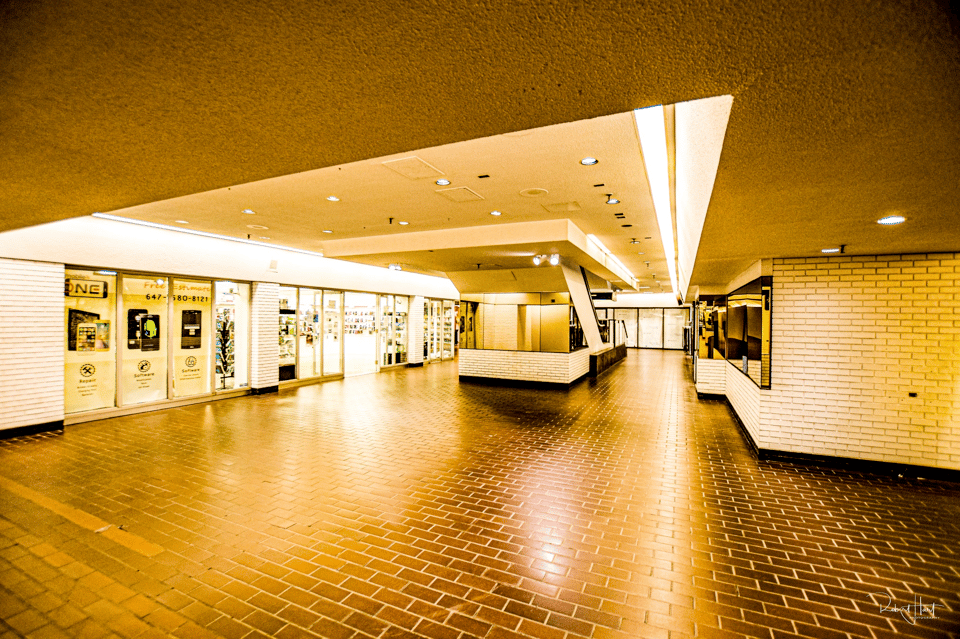 A photo of an old 1980s mall. It is yellow tinted. The floors and walls are all tiled, and there's bright flourescent lighting everywhere